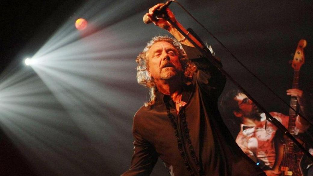 Robert Plant is wearing a black shirt open at the neck. He is holding a microphone with a stand at a 45-degree angle. Behind him are spotlights to the left and a bass player to the right