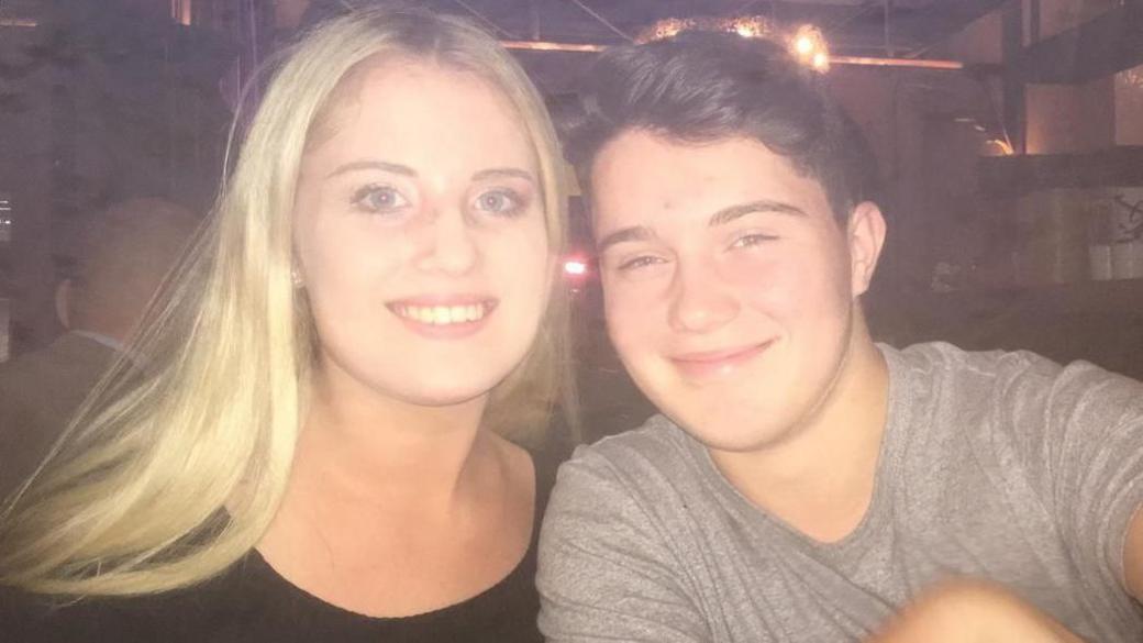 A young man with short, side-parted black hair wearing a grey t-shirt and a young woman in a black top sit next to each other in a dimly lit bar, smiling. The photo looks like it was taken some time ago, and there is a slight haze to the image.