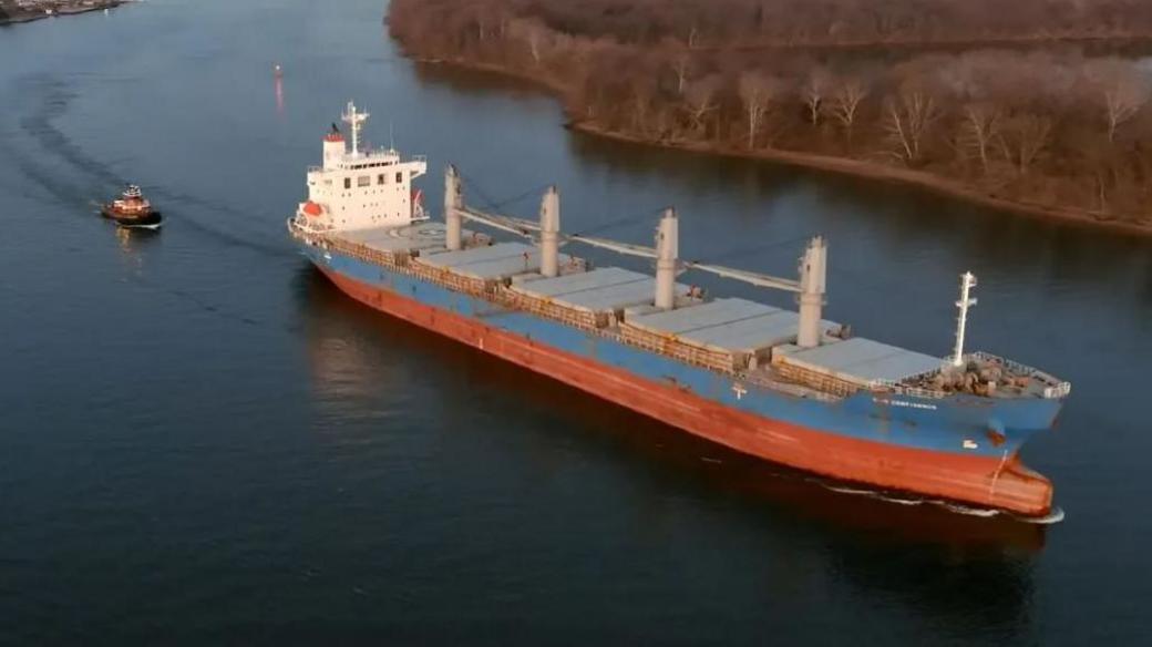 A red and blue cargo ship being led by a tug boat along a river bend at dusk