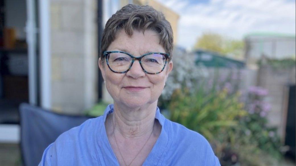 Karen Thompson stood in her garden. She is wearing a blue blouse and has glasses. Behind her there are some out of focus green bushes and pink flowers.