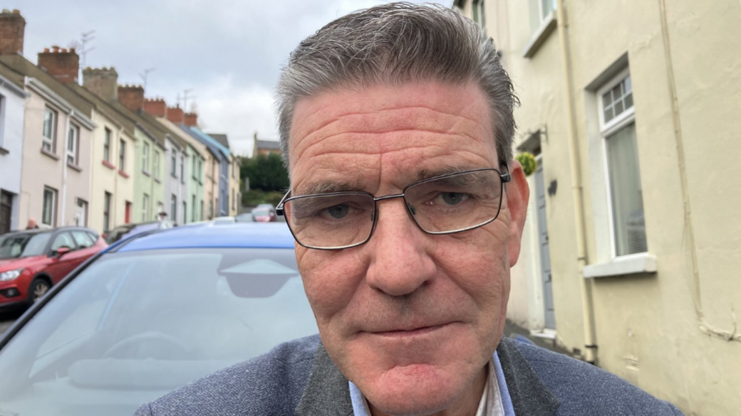Shows a man wearing glasses with a light blue jacket standing in a street with cars in the background.