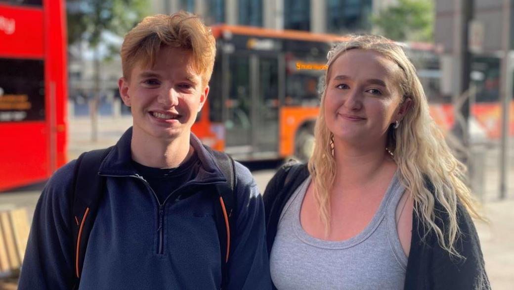 Students Evan Stoddart and Holly Wells, who stand in front of Wood Street near Central Square, Cardiff. Evan (left) has ginger short hair, and wears a dark blue quarter zip fleece and a backpack, while Holly (right) has blonde long hair and wears a grey vest and cardigan. There are buses in the background, which are blurred as the two people, in the foreground are the focus of the picture.