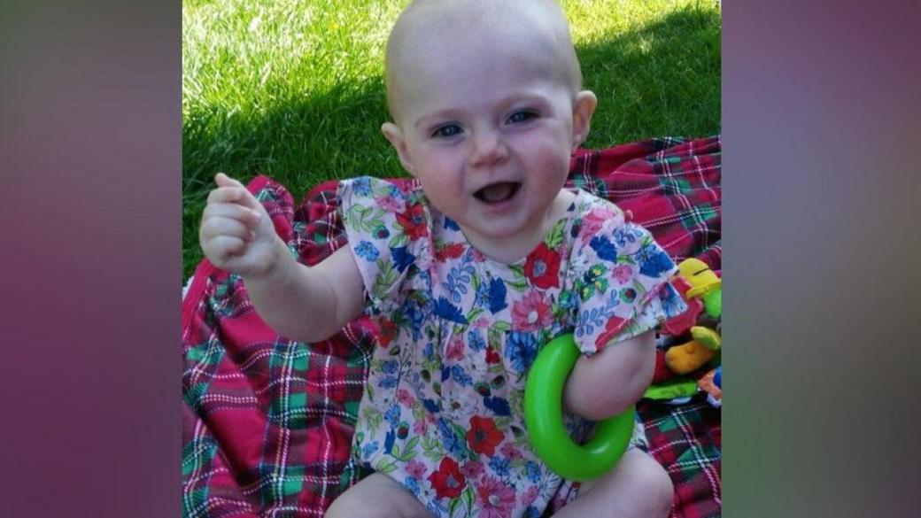 Zoey pictured when she was a baby garden sat on a red and green checked picnic blanket.

She has a blue and pink floral dress on and has a green hoop tucked under her left armpit