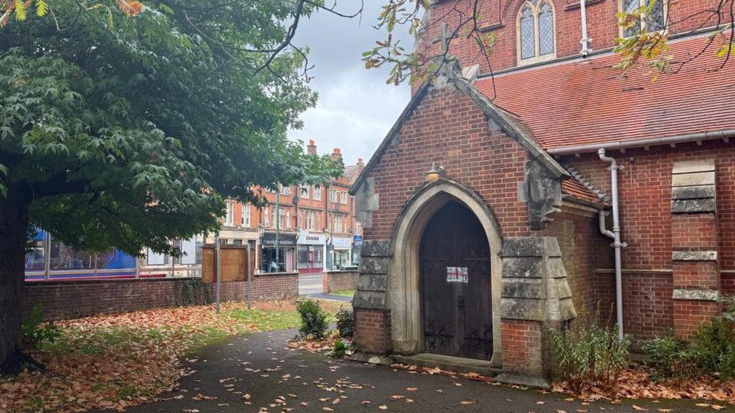 St Luke's Church, Winton. A Victorian brick church with a pitched roof porch and a large double oak door with a crudely-painted George Cross in the centre. The path is Beyond the church wall is a street lined with terraced buildings and shops.