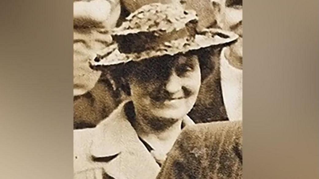 A sepia tone picture of Louisa Dunne. She is wearing a collared coat and wide brimmed hat. She is smiling at the camera in a crowd of people. 