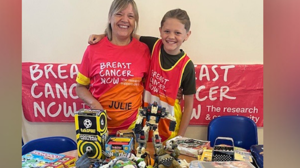 Woman with shoulder-length blonde hair smiling with a red Breast Cancer Now t-shirt. Next to her is a boy with short brown / blonde hair with a black t-shirt and a Breast Cancer Now tabard. There is a red Breast Cancer Now banner behind them on a beige wall. In front of them there is a table with toys on it and there are also two blue chairs. 