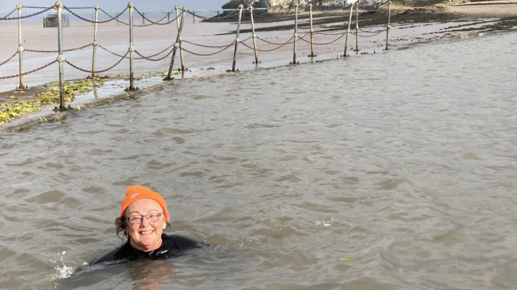 An older woman wearing a black wetsuit and an orange woolly hat, smiling to camera while enjoying a dip in the Clevedon Marine Lake.