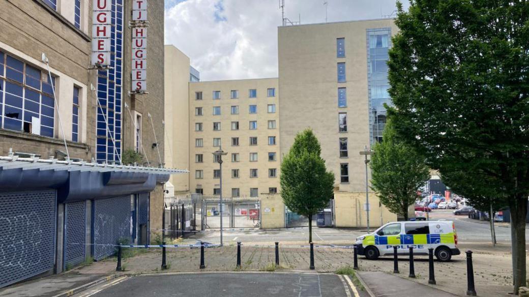 A police van parked next to police tape which is wrapped around a tree in a courtyard with buildings on each side. The department store is on the left with signs which read 'TJ Hughes'