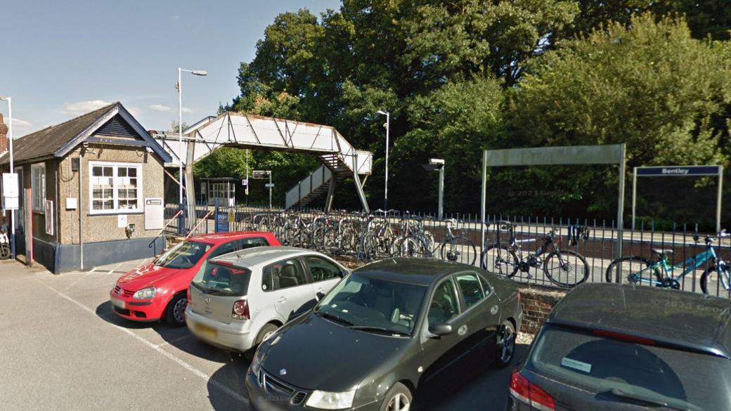 Bentley station, which has a single-story brick building adjacent to a modest parking area, where a mix of vehicles are parked. Beside the car park, a secure bike rack holds numerous bicycles. An elevated footbridge spans the railway tracks, linking both sides of the station.