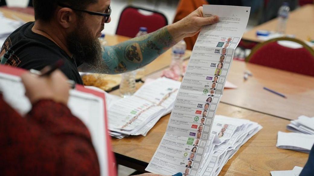 A man with glasses and a long beard and with tattoos up his left arm looks at a ballot paper with around 20 candidates listed on it. 