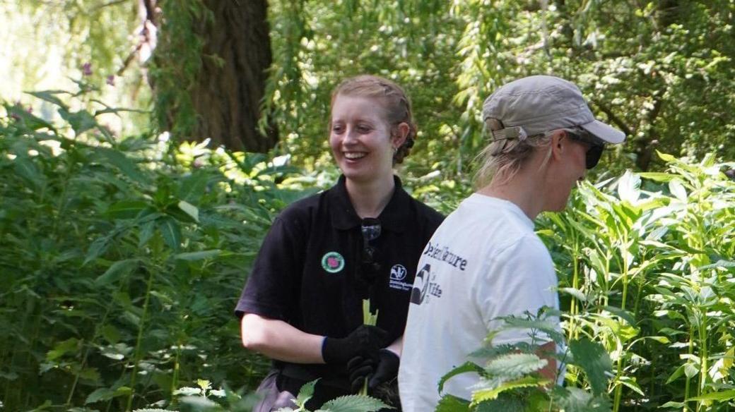 Miriam Lawley, a young woman with ginger hair tied in a bun with a plait across the side of her head. She is wearing black overalls with a Nottinghamshire Wildlife Trust motif depicting a badger, and she is wearing black gloves.