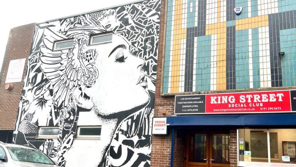Mural covering the side wall of a two-storey building. It is black and white and of a woman in profile, stylised with patterns, including partial faces, around her. On the right is the entrance to King Street Social Club - marked with a sign above the entrance, and a pattern of blue, white, black and beige, tiles above it.