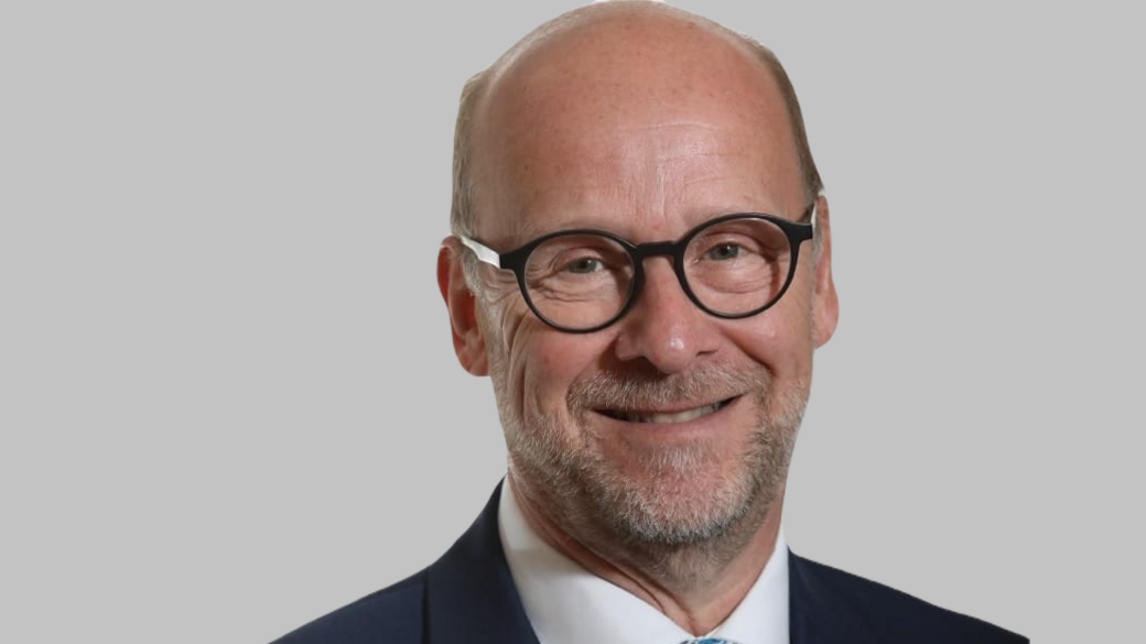 Simon Martin has a bald head, grey stubble and black circular-framed glasses. He is stood in front of a grey wall.