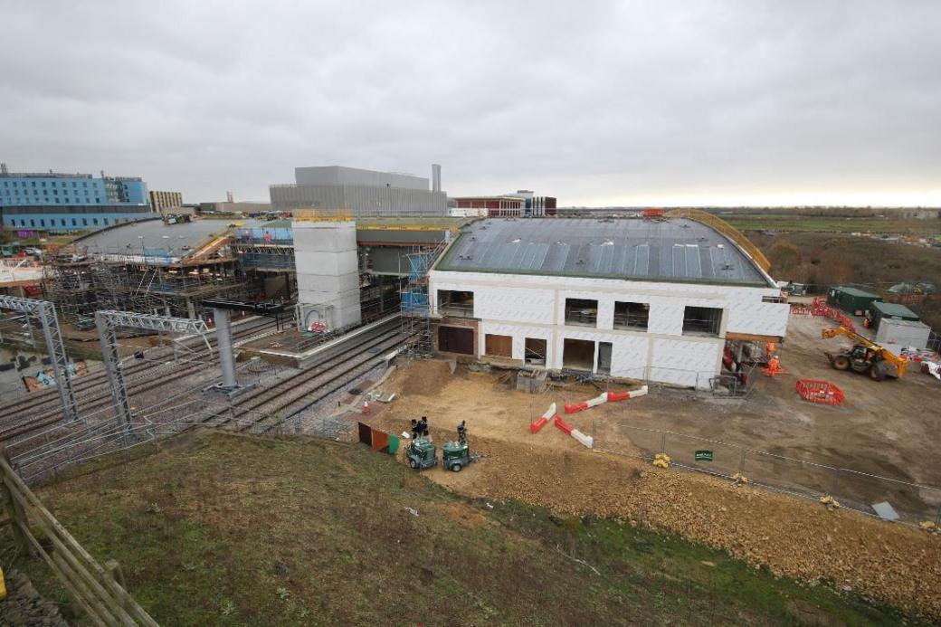 Cambridge South railway station seen from the outside, perhaps a semi-ariel view. Train lines run into the main station undercover, while on the right a new white building is being constructed, with a curved perspex roof. There are a few safety barriers scattered around and a JCB in the distance. Behind the station are what look like offices/hotels and industrial buildings.