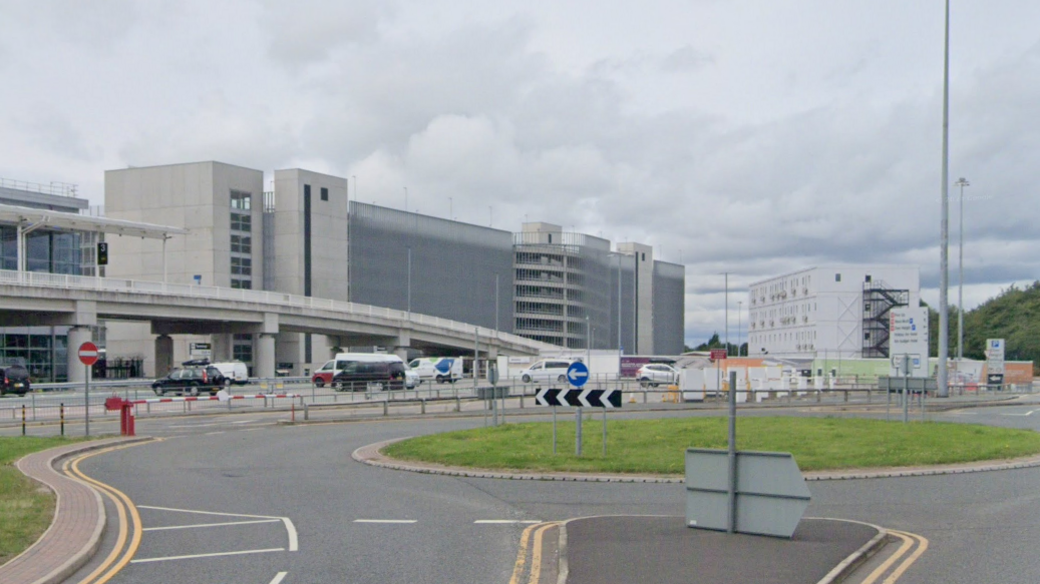 A shot of Manchester Airport showing a car park and terminal building behind a roundabout