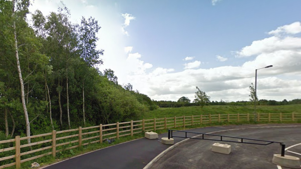 Trees to the left of the picture, with a field to the back and a path and road to the foreground, with a metal and concrete barrier in place