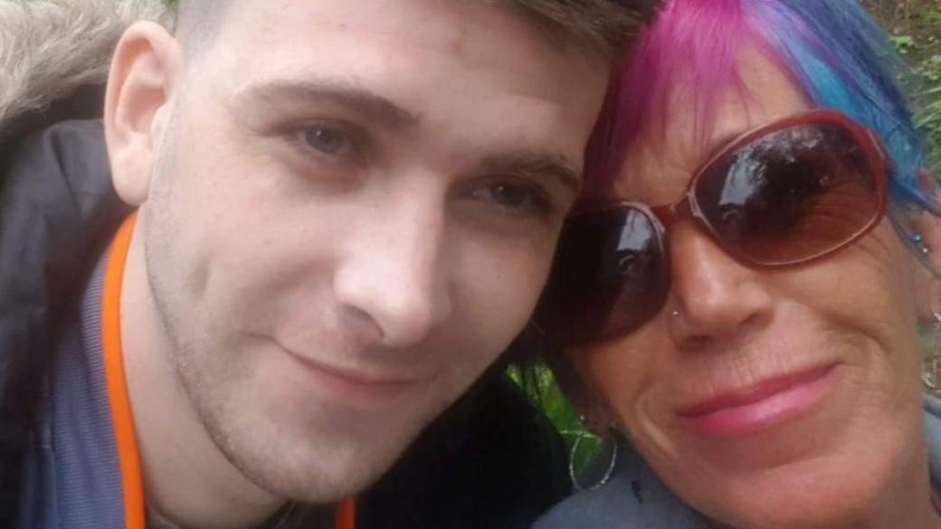 A selfie picture of James with his mother Jayne. They are both quite close to the camera, looking directly at it. She is wearing sunglasses and has hair which is coloured purple and blue. He is wearing a parka jacket.