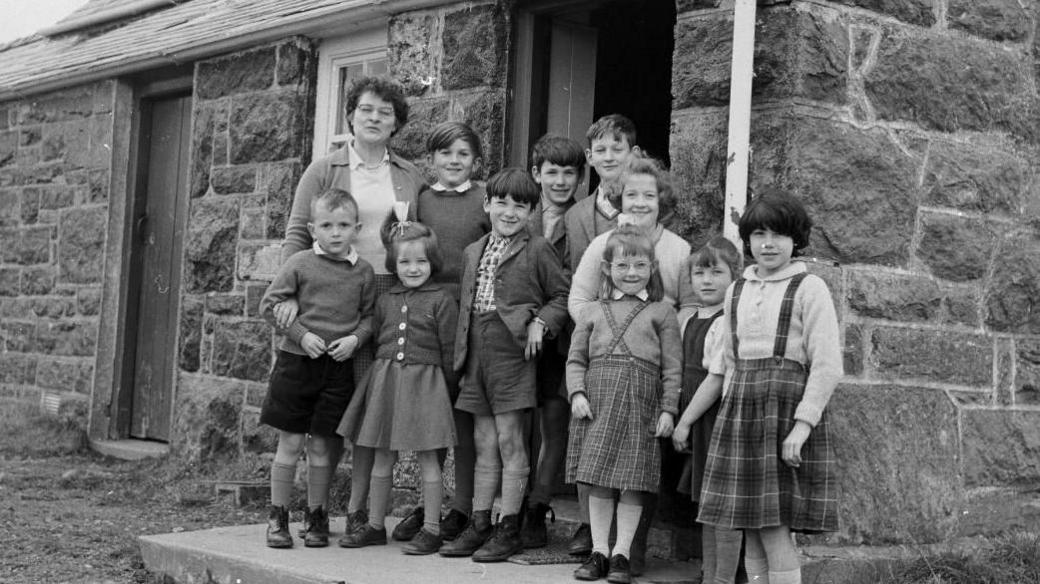 Black and white photograph showing very young school pupils outside the school of Capel Celyn. Ten young school pupils can be seen huddled on the school steps. The boys were shorts, jumpers and shirts. While the girls were tartan skirts and cardigans. Their teacher stands tall at the back of the group, they all smile at the camera. 