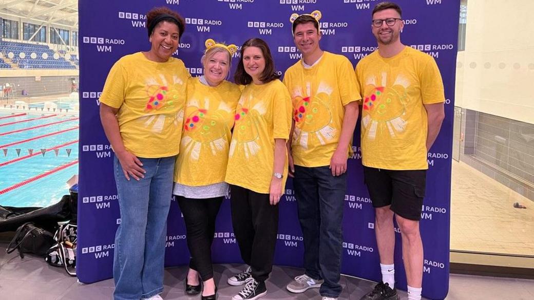 Five people wear yellow t-shirts in front of a purple board with BBC Radio WM branding. The pool is visible in the background to the left.