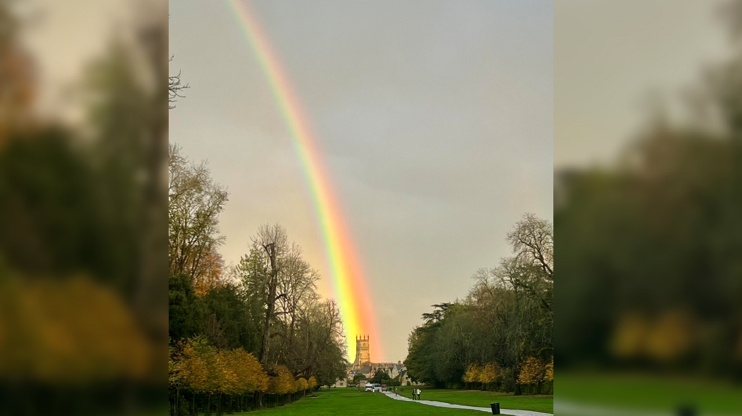 A bright rainbow shoots down the sky above Cirencester Park, which is surrounded by trees. At the bottom of a long path down the lawn lies St John Baptist Cirencester church, where the rainbow ends.
