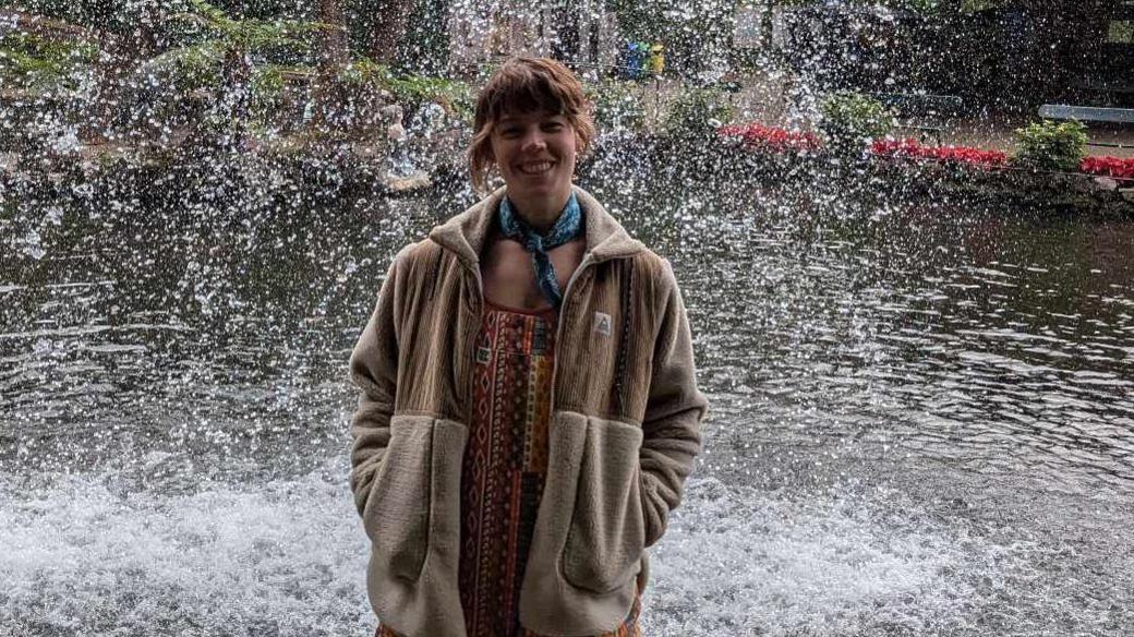 A woman stands in front of a fountain, smiling at the camera.