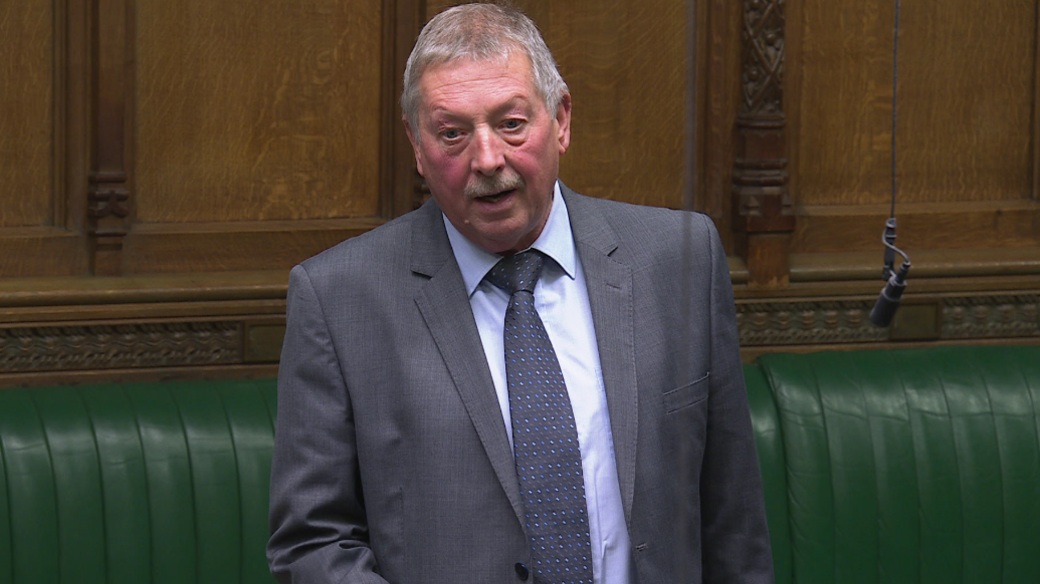 Sammy Wilson speaking in the House of Commons. He has a grey suit on with a blue polka dot tie and blue shirt. He has gray balding hair and a gray moustache. Behind him are green leather seats.