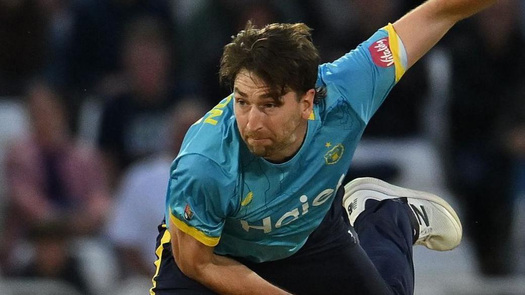 Bowler Richard Gleeson, wearing a blue shirt and navy trousers, watches after bowling a ball while playing cricket for Warwickshire Bears