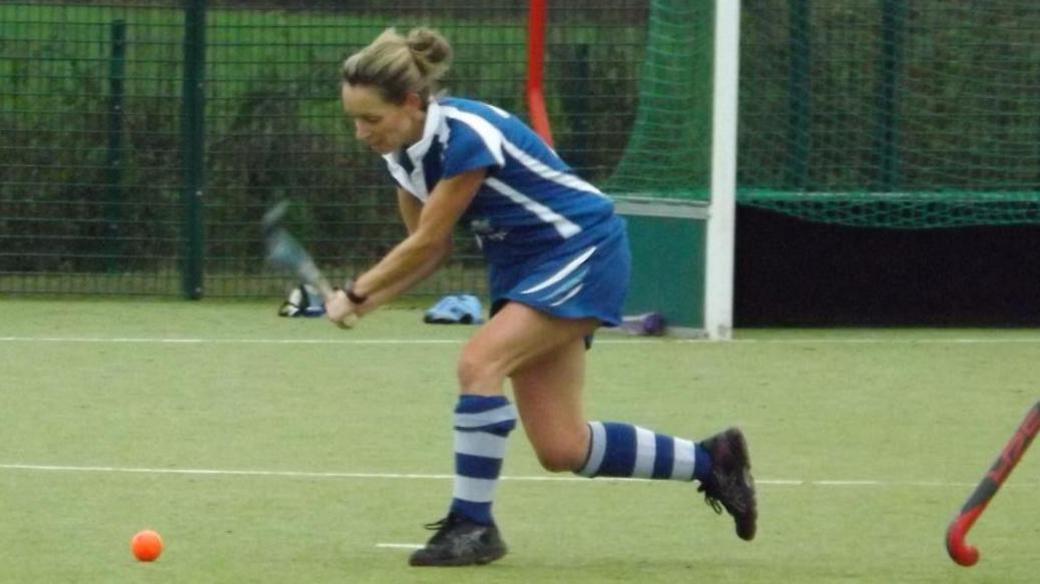 Hockey player Claerwen Render about to strike an orange ball with her hockey stick on an all weather pitch. She is wearing a blue hockey kit with blue and white striped socks.