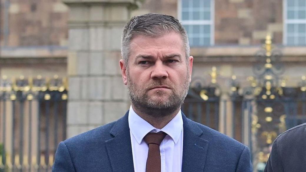 Colin McGrath, a man with short, greying hair and matching beard, standing outside Hillsborough Castle during a previous press event. He is wearing a navy suit, a white shirt and a brown tie. The Georgian-era brown sandstone building and its black and gold gates are in the background.