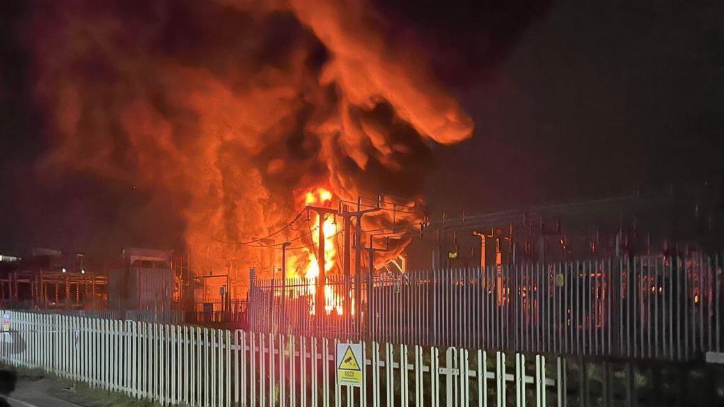 Black smoke and large yellow flames billow into the night sky in between power cables at the electrical substation. Rows of metal fences can be seen in the foreground