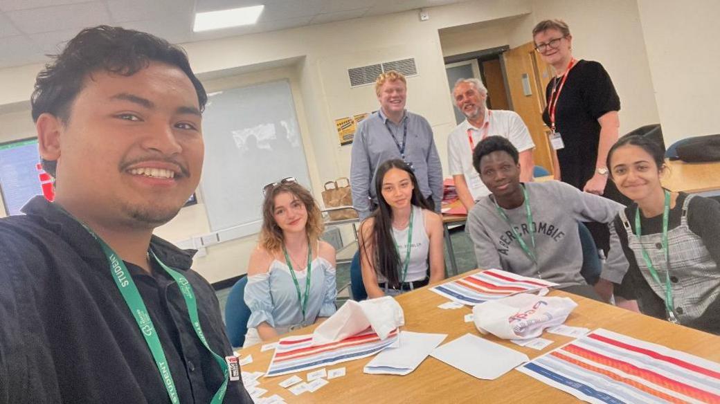 A group of five students in a room, with one standing up, with paper work in front of them and three adults standing behind them. They are all wearing lanyards, smiling and looking at the camera. 