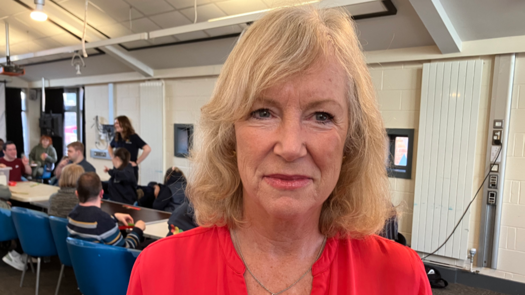 A woman with light coloured hair wearing a light red top and a necklace smiles to the camera. She is an office with people sat at a table behind her.