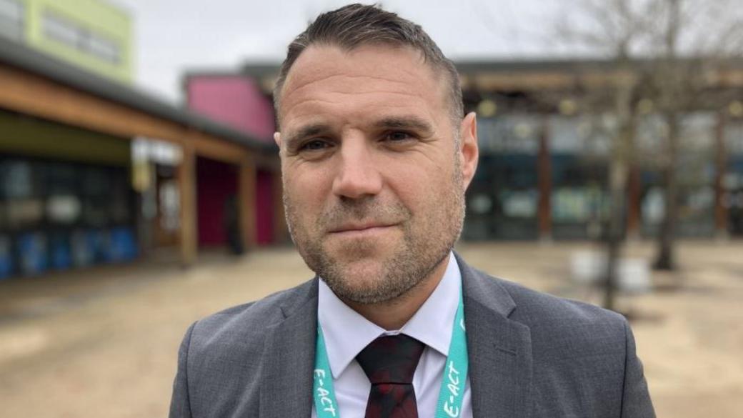 Headteacher Guy Swallow stood outside Merchants' Academy Secondary School. He is wearing a grey suit, white shirt and a tie. The school building behind him is blurred. 