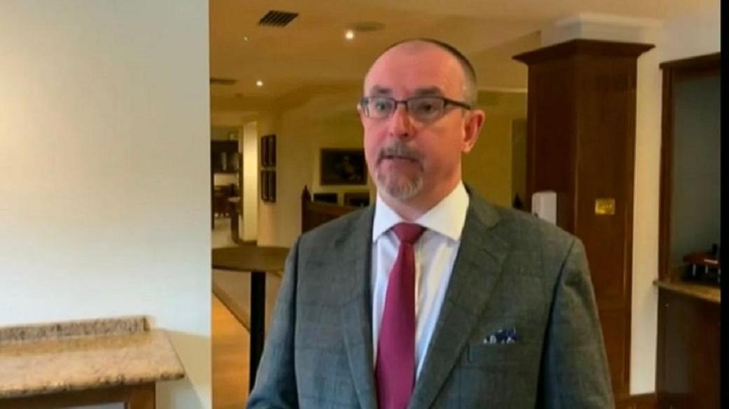 Andrew Hollett with short dark hair with a slight beard and round glasses wearing a grey jacket, red tie and white shirt. He is standing in a hotel- a wood pillar is visible behind him to the right and a bar to the left.