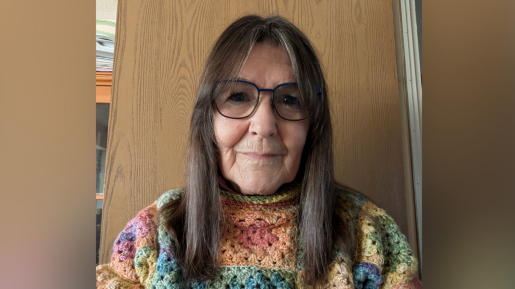 A head and shoulders image of Penny Dack. She is wearing a multi-coloured jumper and glasses and standing in front of a wooden backdrop. She is looking directly into the camera.