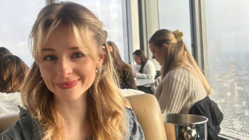 Eve Dulcie sits inside a in a rooftop restaurant with a skyline in the background. She smiles at the camera. She has blonde mid length hair, which is half pulled back from her face with her bangs loose in her face.
