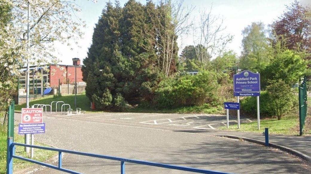 The entrance Ashfield Park Primary School has an entrance sign and a speed limit reminder, with trees, shrubs and areas of grass.