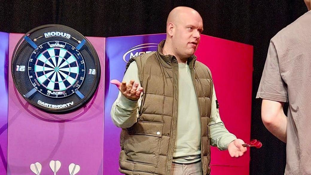 Michael Van Gerwen standing next to a dart board with darts in his hand at Nescot in Epsom