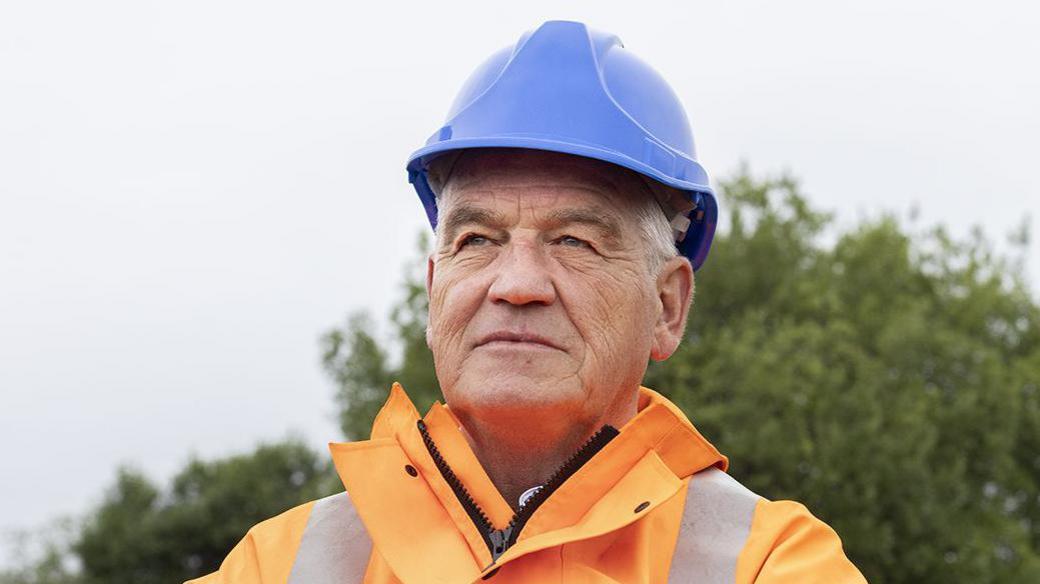 Gateshead Council leader Martin Gannon at the Leamside Line, wearing a blue hard hat and orange hi-vis jacket. He is staring into the distance.