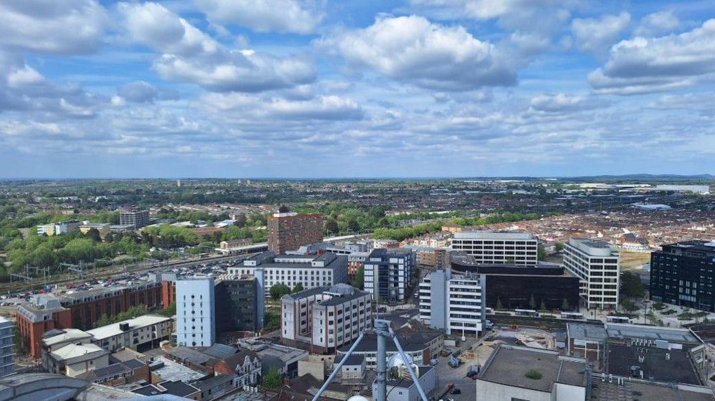 Birds-eye view of Swindon's centre, railway line and trees in the background. Lots of high-up buildings.