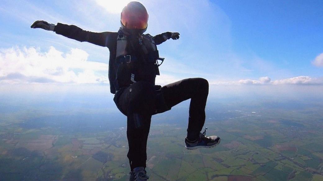 Michael Oakes midway through a skydive, high above the landscape. He is in a seated position with his arms and legs outstretched. The sun is behind him so, with the exception of his reflective orange helmet visor, he is almost silhouetted against the blue sky.