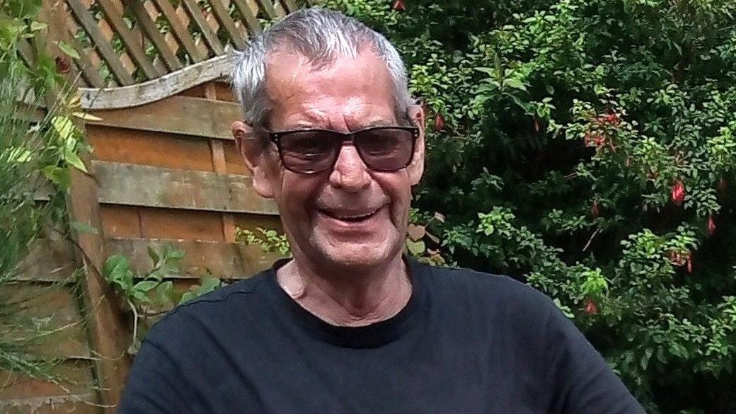 Abraham Torrance smiles into the camera while sitting in a garden. He has short grey hair and is wearing a black t-shirt and dark sunglasses. Behind him is a fence and luscious greenery.