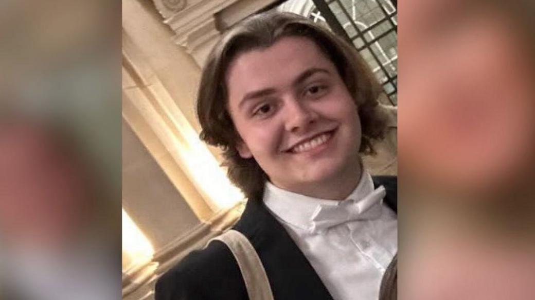 Connor is smiling at the camera, wearing a white shirt and bow tie, and black dinner jacket. There's a beige bag strap over his shoulder.