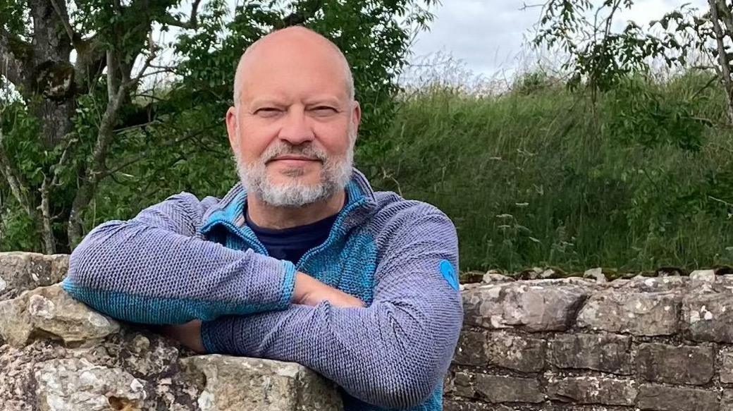 A head-and-shoulder image of Stephen Cooper. He is outdoors and leaning on a drystone wall. He is bold, with white trimmed beard and moustache. He is wearing a purple and blue jumper and he is slightly smiling.