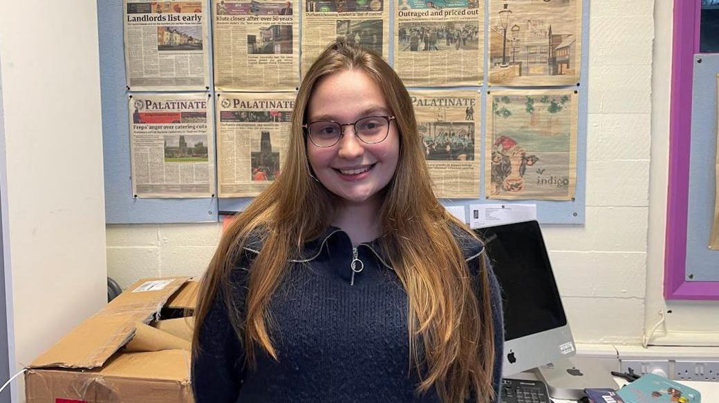 Student Lily Gershon, standing in her university newspaper office with a computer and copies of the university newspaper behind her. She has long blonde hair, is wearing glasses and smiling at the camera.