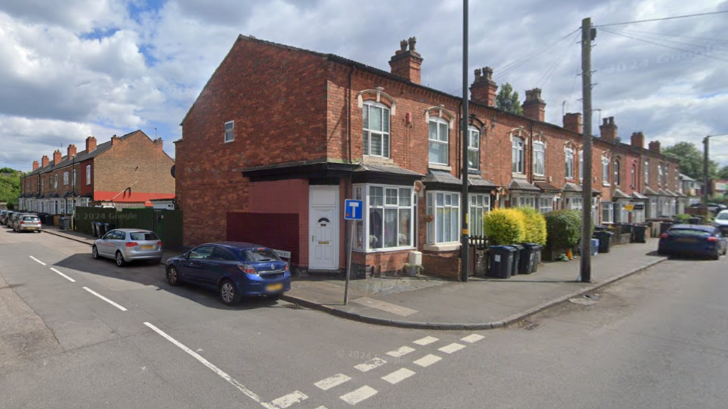 The junction of two roads, with a row of terraced houses stretching back from the camera, and cars parked to the side of the end terrace.