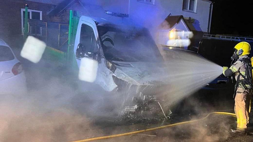 Firefighter spraying water on a damaged white vehicle amid smoke