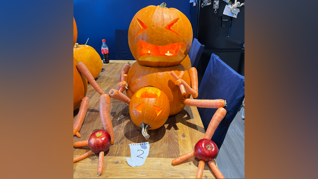 A carved pumpkin "head" sits on another slightly bigger pumpkin "body", and appears to be giving birth to a smaller carved pumpkin. Carrots make up the arms and legs. Each foot is one red apple. The creation, which has two candles in its mouth, is sitting on a wooden dining table.