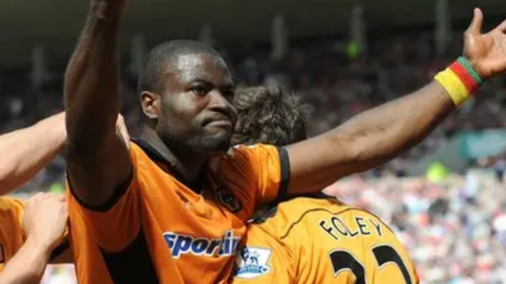 Footballer in an orange short-sleeved Wolves shirt with a wristband showing the yellow, red and green colours of Cameroon accepts the acclaim of supporters after scoring a goal