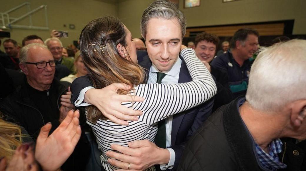 Simon Harris who has greying hair parted to the side and is wearing a dark side is hugged by his wife who has long brown hair and is wearing a striped top at an election count centre